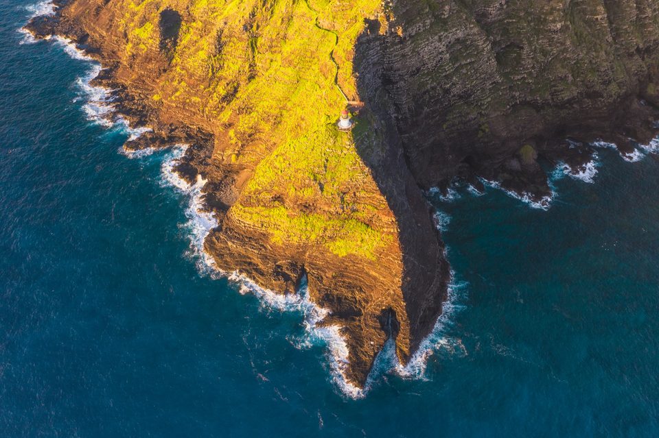 Aerial sunrise view of the Makapuʻu cliffs and lighthouse, one side lit gold and the other in shadow above deep blue water