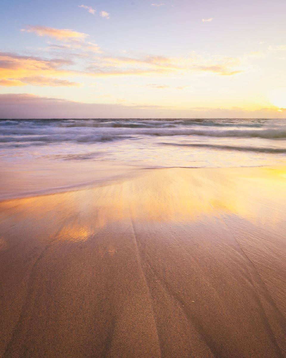 Black sand beach at sunset, fine lines drawn in the wet sand by a receding wave under a pastel sky