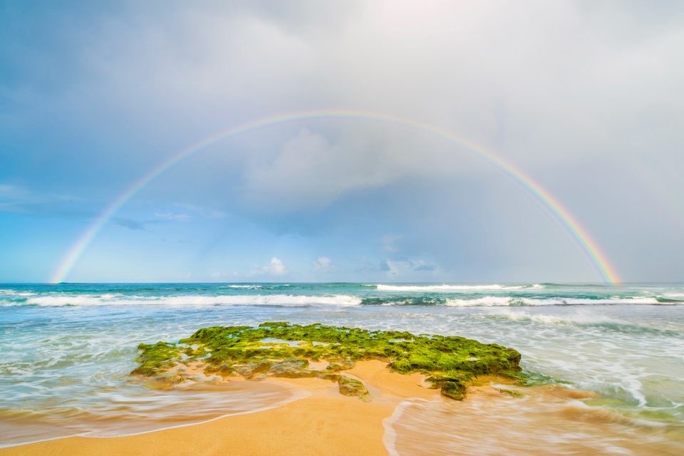 A full rainbow arching over the North Shore surf, a moss-covered rock on the sand in front