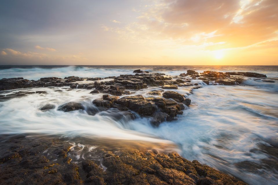 Long exposure of waves washing over lava rock at South Point on Hawaiʻi Island, the sun low over the ocean