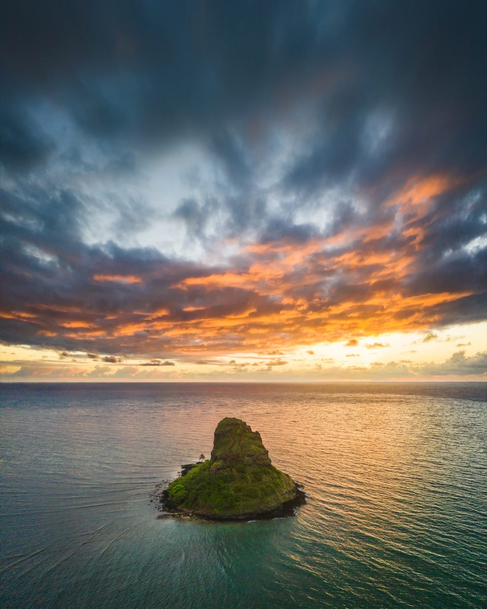 A lone islet in a calm sea under dramatic orange sunrise clouds
