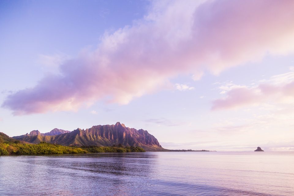 The Kaʻaʻawa ridge glowing pink at sunrise, mirrored in calm water seen from Waikāne