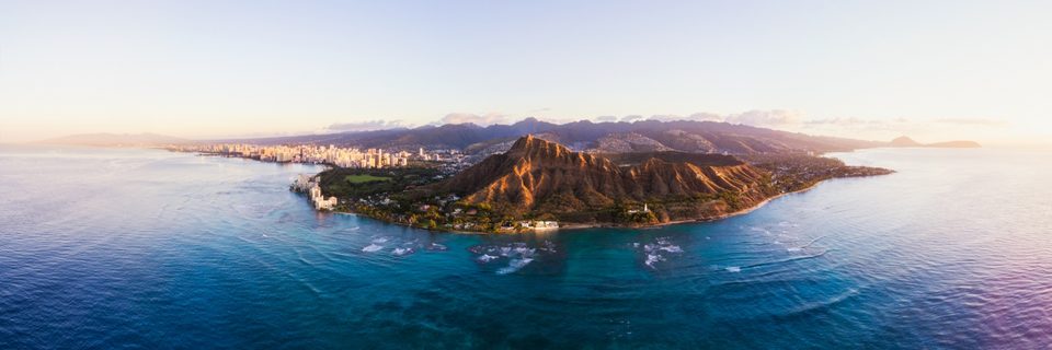 Aerial panorama of Diamond Head and Waikīkī at sunrise, gold light on the crater and turquoise reef below
