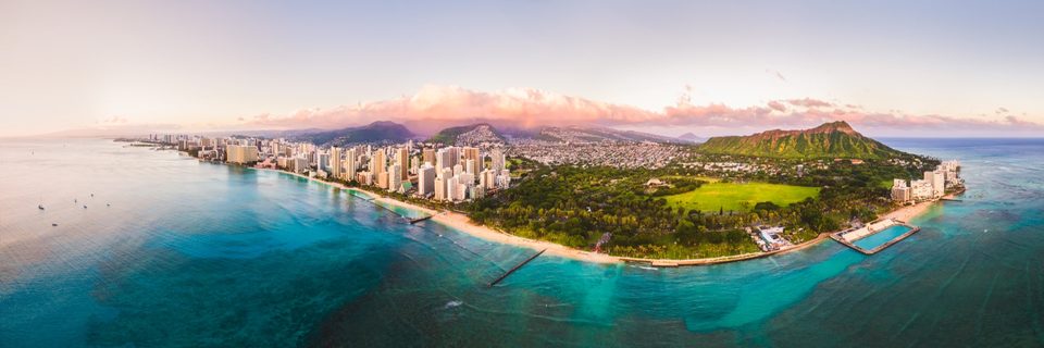 Aerial panorama of Waikīkī and Diamond Head at sunset, pink clouds over the city and turquoise reef below
